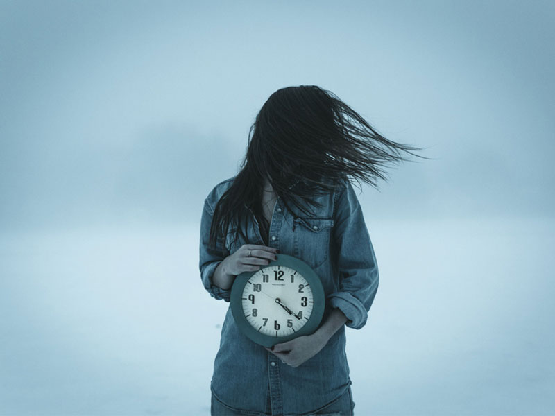 γυναίκα με τζιν πουκάμισο και ανακατεμένα μαλλιά κρατά ένα ρολόι και έχει κατάθλιψη