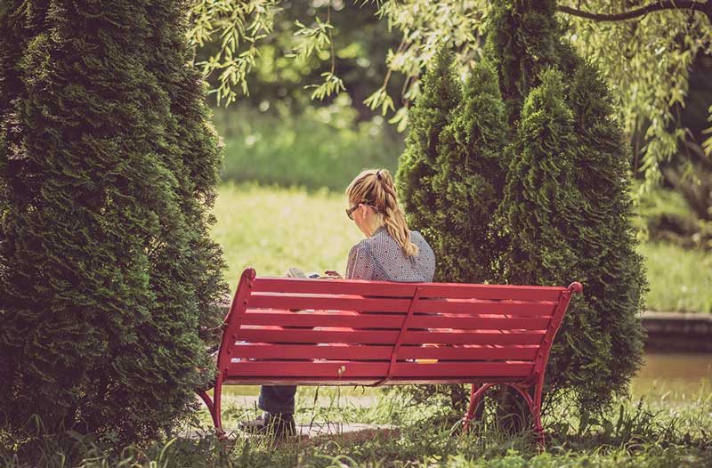 γυναίκα κάθεται μόνη σε κόκκινο παγκάκι στο πάρκο διαβάζοντας βιβλίο έχει καλές σχέσεις αφού έχει καλή σχέση με τον εαυτό της