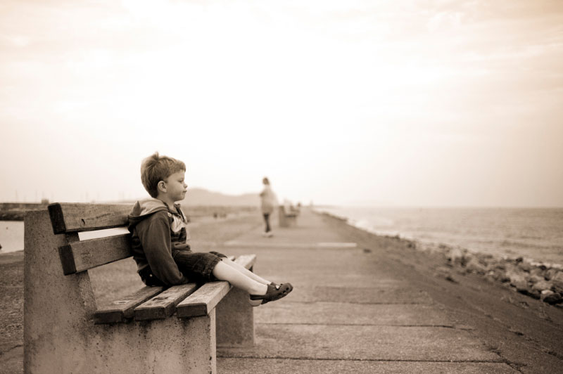 παιδί που κάθεται μόνο του και παραμελημένο σε παγκάκι κοντά στη θάλασσα