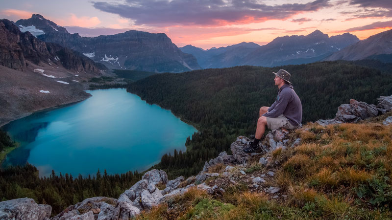 άντρας κάθεται σε ένα βράχο και κοιτάζει πράσινο τοπίο με λίμνη