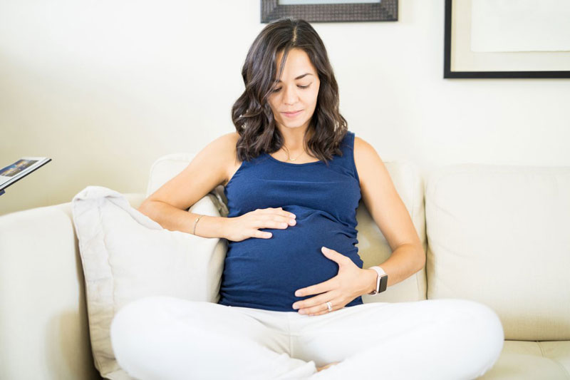 έγκυος γυναίκα καθισμένη σε έναν καναπέ χαϊδεύει τη κοιλιά της