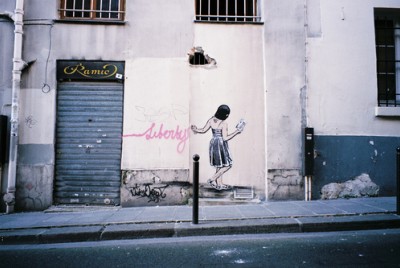 γκράφιτι σε φθαρμένο τοίχο που απεικονίζει μία κοπέλα που γράφει Liberty με ροζ γράμματα