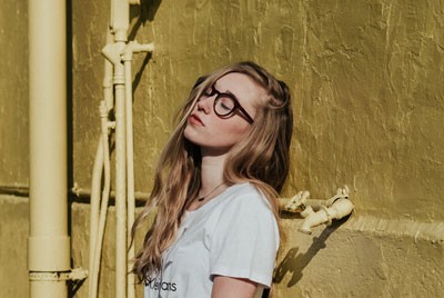 κορίτσι με γυαλιά και λευκό μπλουζάκι που στέκεται με την πλάτη σε ένα τοίχο και έχει κλειστά μάτια