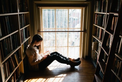 κοπέλα κάθεται ανάμεσα σε δυο βιβλιοθήκες και διαβάζει ένα βιβλίο