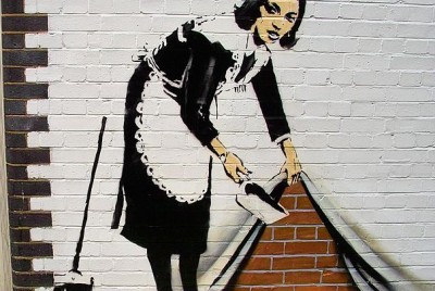 γκράφιτι γυναίκας ντυμένης καμαριέρας σε τοίχο που κρύβει κάτω από το σεντόνι τα σκουπίδια