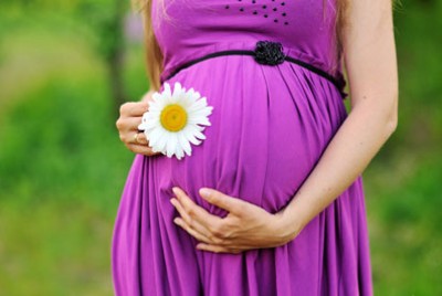 έγκυος που πιάνει την κοιλιά της φορώντας ένα μωβ φόρεμα και κρατώντας ένα λουλούδι