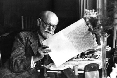 o Sigmund Freud στο γραφείο του σε ασπρόμαυρη φωτογραφία να σηκώνει χαρτιά που έχει γράψει