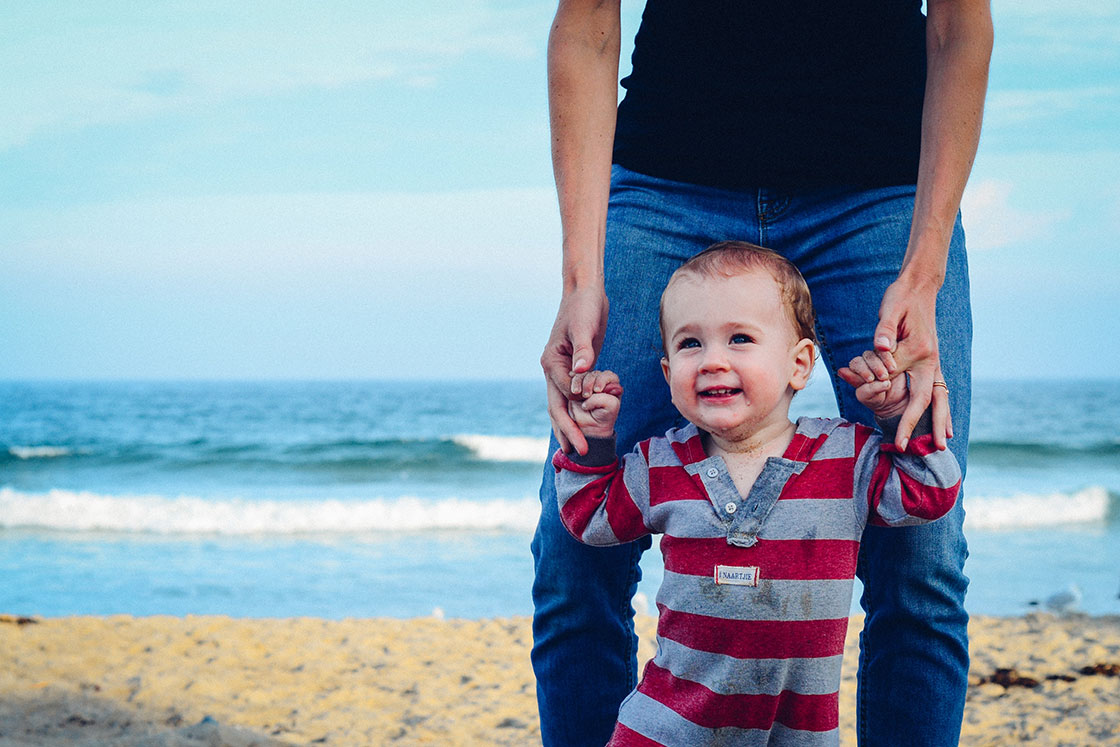 μωρό που χαμογελάει καθώς το κρατάει ο πατέρας του σε μία παραλία με φόντο τη θάλασσα