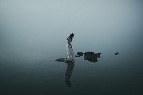 γυναίκα με λευκό φόρεμα στέκεται σε ένα βράχο μέσα στη θάλασσα και απεικονίζει την κατάθλιψη και τα μυστικά της