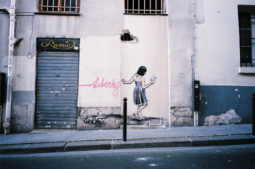 γκράφιτι σε φθαρμένο τοίχο που απεικονίζει μία κοπέλα που γράφει Liberty με ροζ γράμματα