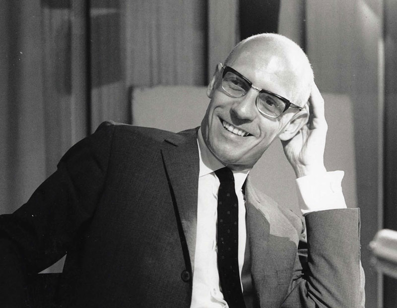 o Michel Foucault με γυαλιά χαμογελάει πιάνοντας το κεφάλι του