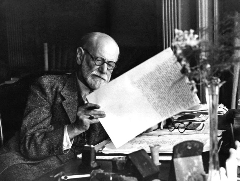 o Sigmund Freud στο γραφείο του σε ασπρόμαυρη φωτογραφία να σηκώνει χαρτιά που έχει γράψει