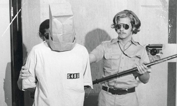 φύλακας με στολή σπρώχνει με το γκλομπ κρατούμενο με κουκούλα στο κεφάλι σε ασπρόμαυρη φωτογραφία