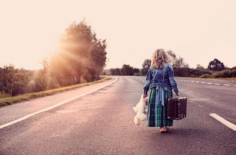 κοριτσάκι που κρατά βαλίτσα και ένα αρκουδάκι περπατά ξυπόλητο σε ένα δρόμο
