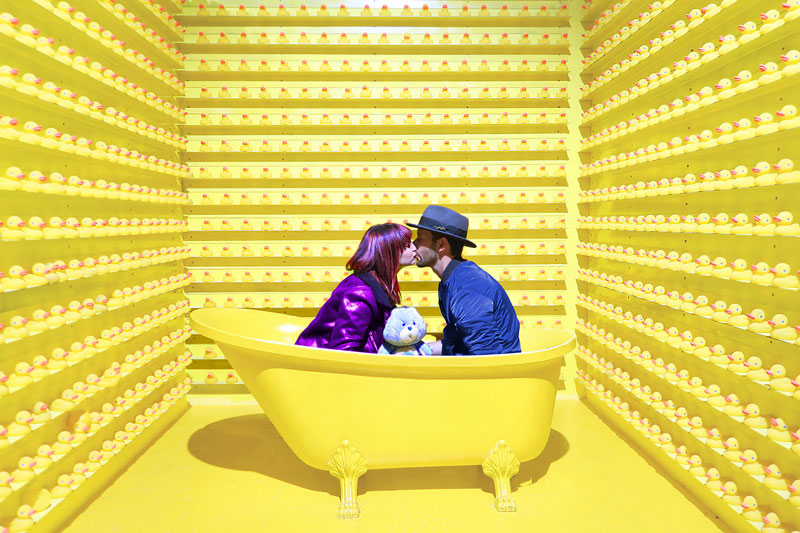 παράξενο ζευγάρι που βρίσκεται σε μία μπανιέρα με ρούχα και φιλιέται σε ένα κίτρινο δωμάτιο