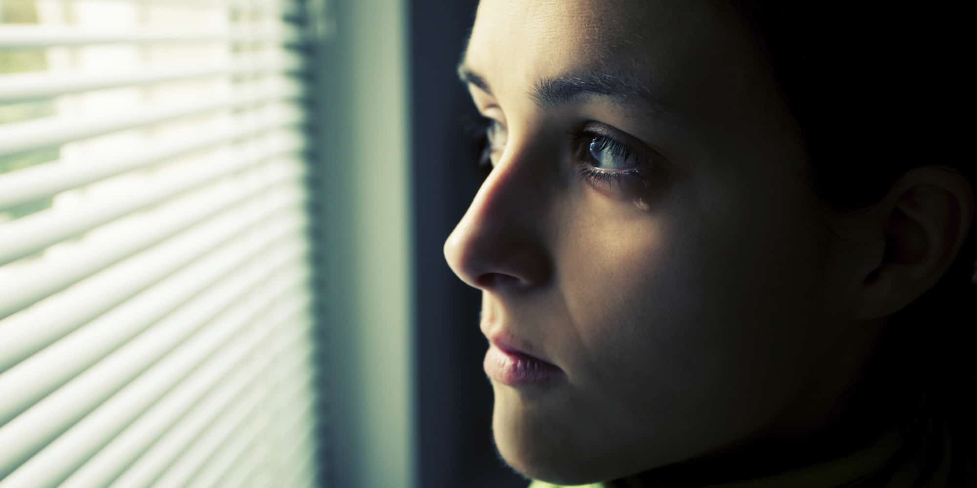 γυναίκα κοιτάει μέσα από τις περσίδες ενός παραθύρου με ένα δάκρυ να κυλάει
