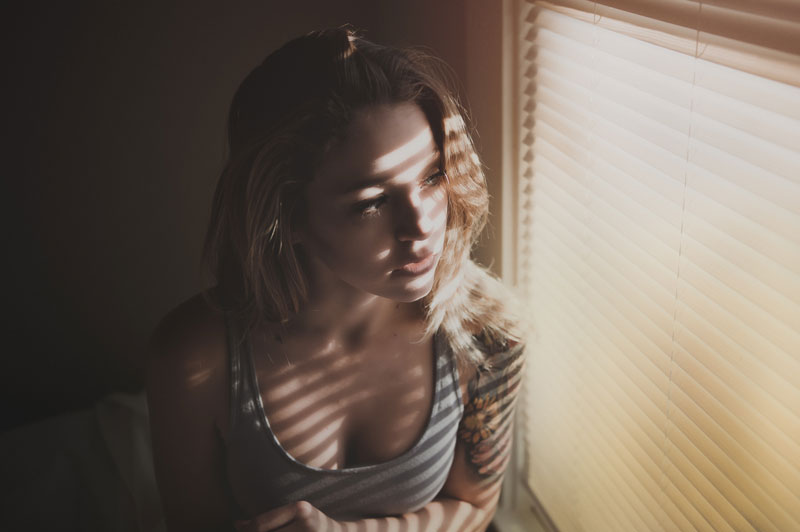 γυναίκα σε σκοτεινό δωμάτιο δίπλα σε παράθυρο που φωτίζεται από τον ήλιο